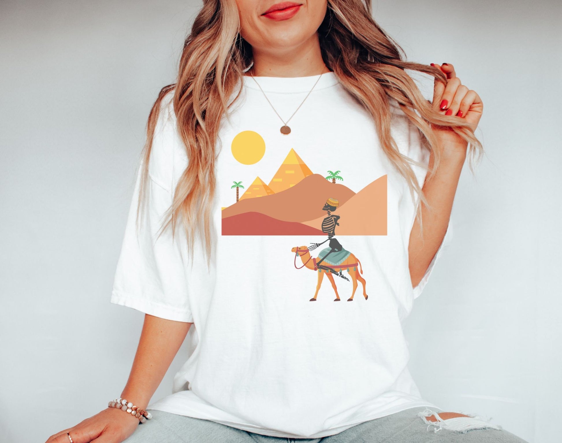 A woman wearing a cute, vintage white colored shirt with a skeleton wearing an Egyptian hat riding a brown camel through the desert, the Egyptian pyramids in the distance behind sand dunes and palm trees with the sun high in the sky.
