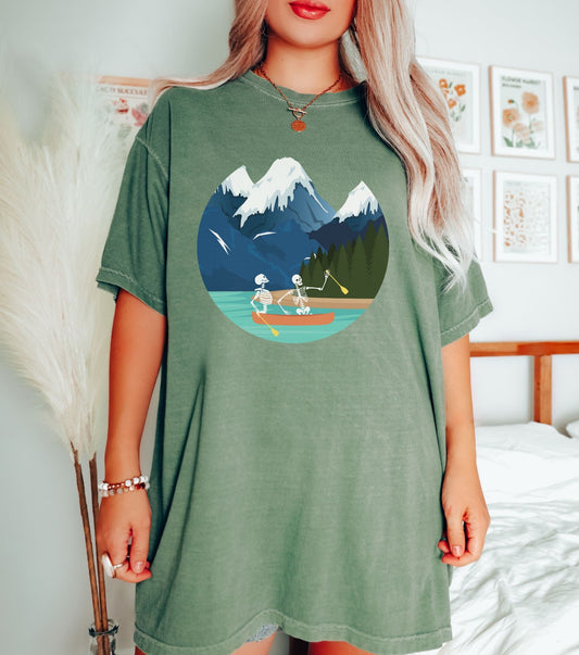 A woman wearing a cute, vintage moss colored shirt with a picture of a skeleton couple rowing a canoe with yellow paddles through a river with snow-capped mountains in the background.