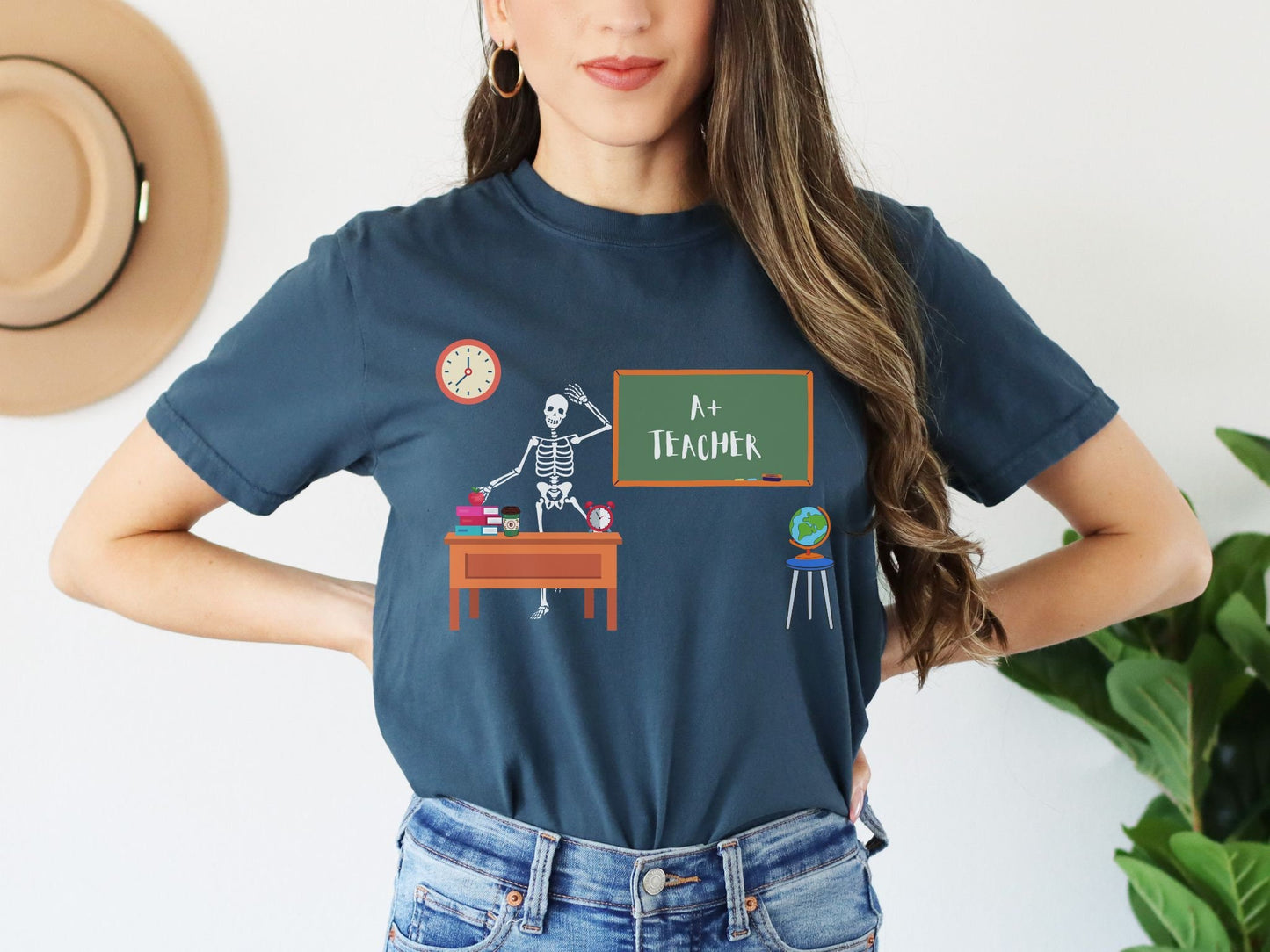 A woman wearing a cute, vintage navy blue colored shirt with a skeleton teacher waving from their desk in front of the green chalkboard which has written A+ Teacher on it. On the teacher’s desk are colorful books, an apple, a coffee, and a clock.