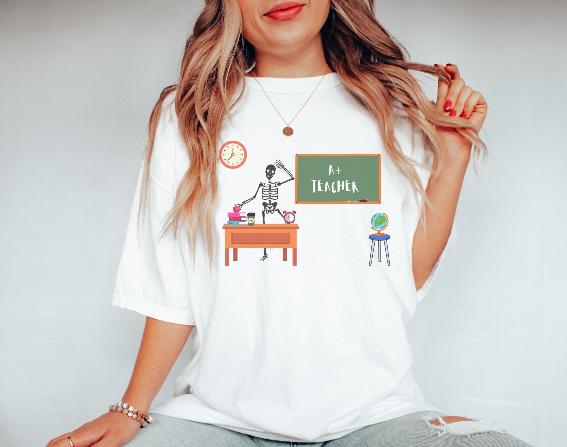 A woman wearing a cute, vintage white colored shirt with a skeleton teacher waving from their desk in front of the green chalkboard which has written A+ Teacher on it. On the teacher’s desk are colorful books, an apple, a coffee, and a clock.