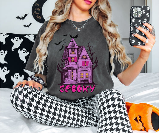 A woman wearing a cute, vintage pepper colored Comfort Colors T-shirt with the text Spooky in purple across the bottom. Above is a Purple and pink haunted house with dark trees in the background with black bats flying above.