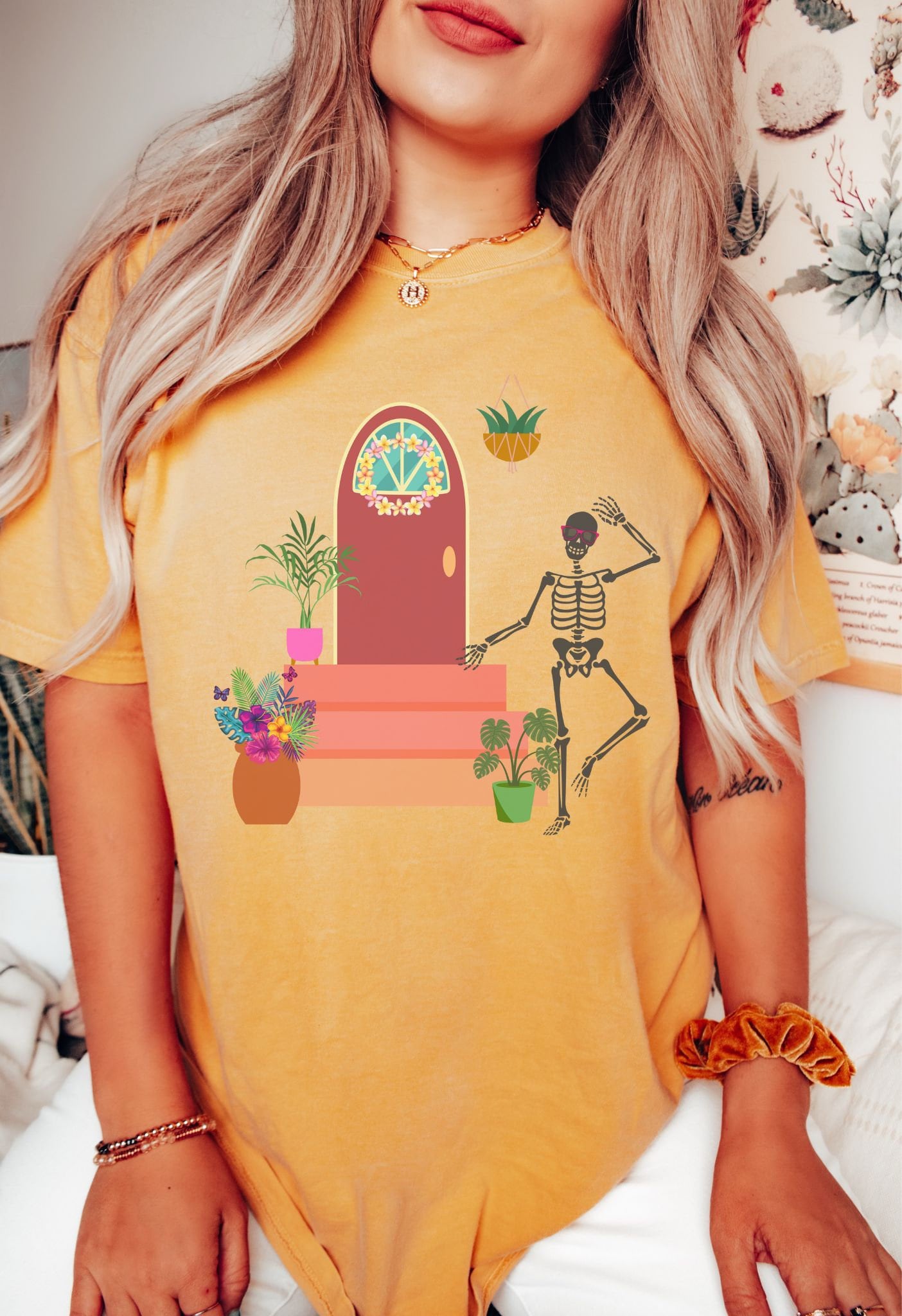A woman wearing a cute, vintage mustard colored shirt with a skeleton wearing red sunglasses walking out the front door of their house. The front stoop has colorful plants and flowers and the front door has a pink and yellow flower wreath hanging.