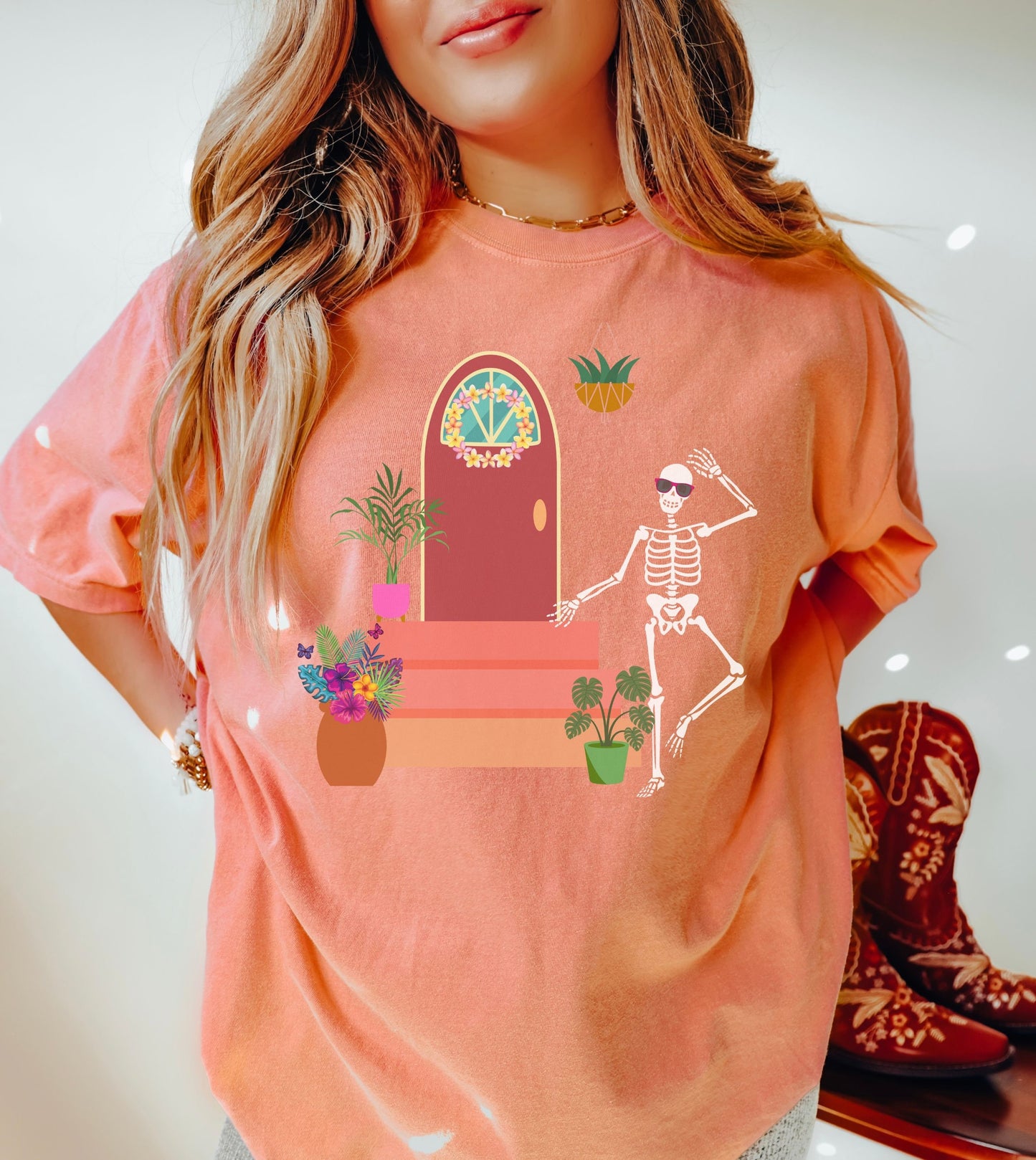 A woman wearing a cute, vintage terracotta colored shirt with a skeleton wearing red sunglasses walking out the front door of their house. The front stoop has colorful plants and flowers and the front door has a pink and yellow flower wreath hanging.