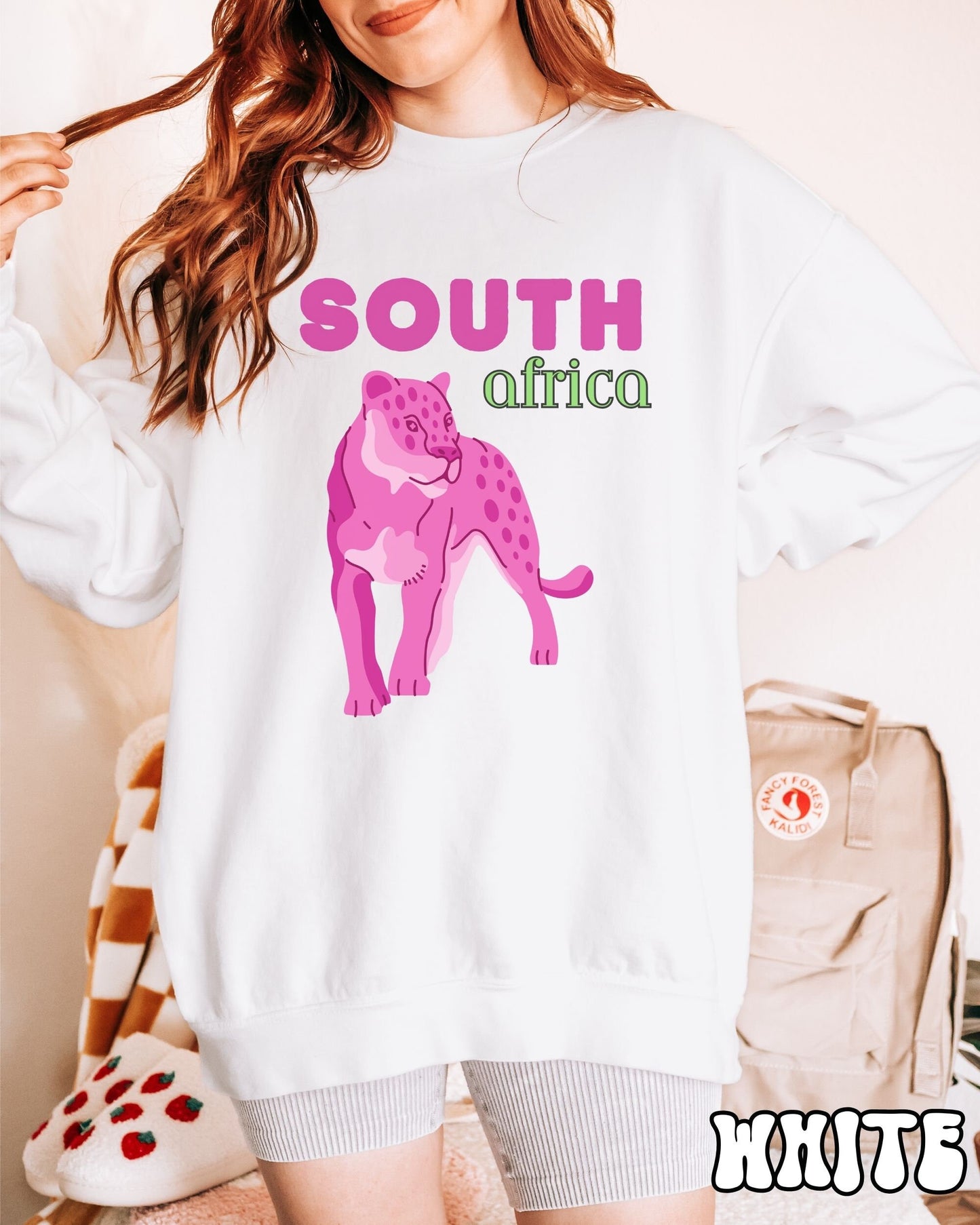 A woman wearing a vintage, white colored sweatshirt with the text South Africa in pink and green font, respectively, and a pink tiger underneath on the prowl.