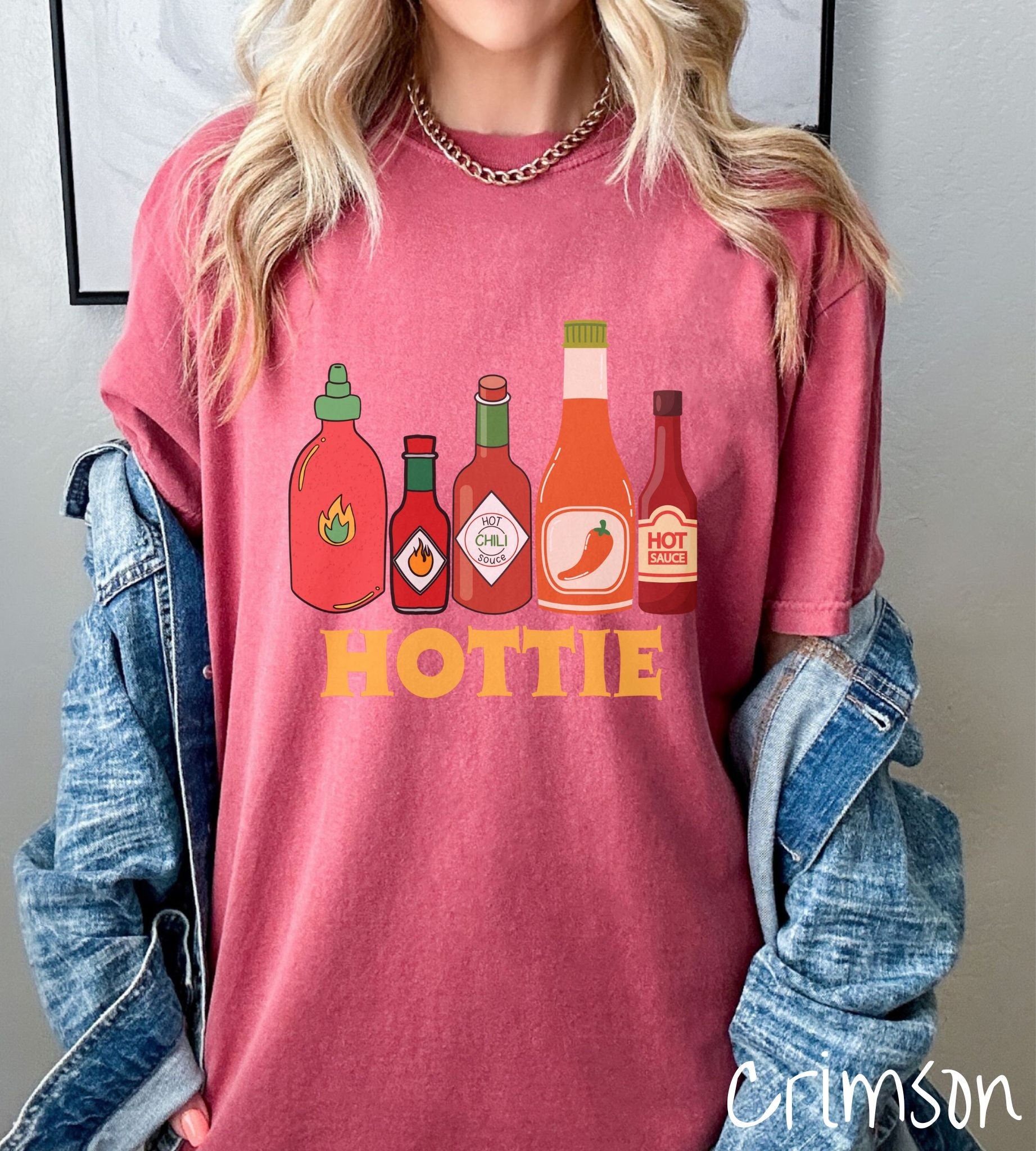 A woman wearing a vintage, crimson colored shirt with red, orange, and dark red bottles of spicy and hot sauces with fire and hot pepper pictures on the labels and the text Hottie in thick red font underneath the bottles and centered on the shirt.