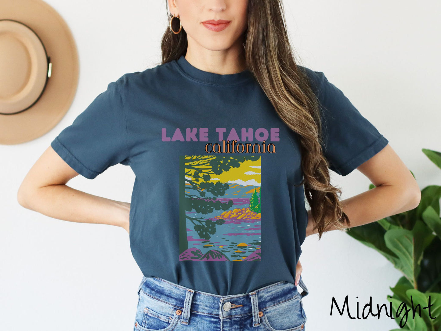 A woman wearing a vintage, midnight colored shirt with the text Lake Tahoe at the top in purple and orange font, respectively, and below is a beautiful picture of the ocean through the trees and rolling hills in the background.