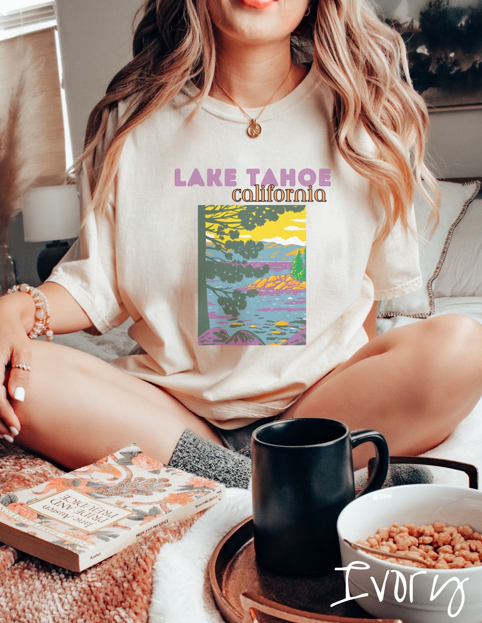 A woman wearing a vintage, ivory colored shirt with the text Lake Tahoe at the top in purple and orange font, respectively, and below is a beautiful picture of the ocean through the trees and rolling hills in the background.