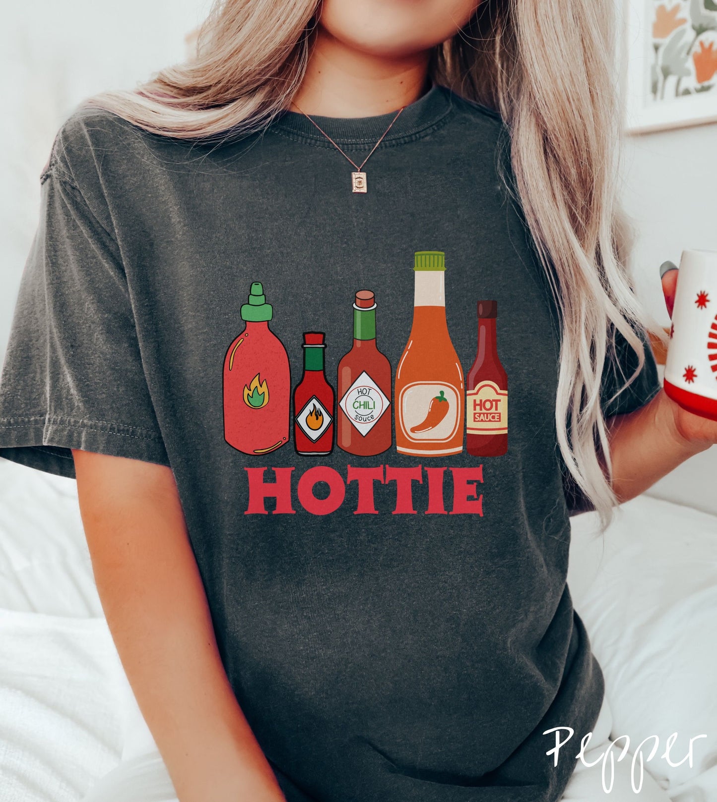 A woman wearing a vintage, pepper colored shirt with red, orange, and dark red bottles of spicy and hot sauces with fire and hot pepper pictures on the labels and the text Hottie in thick red font underneath the bottles and centered on the shirt.