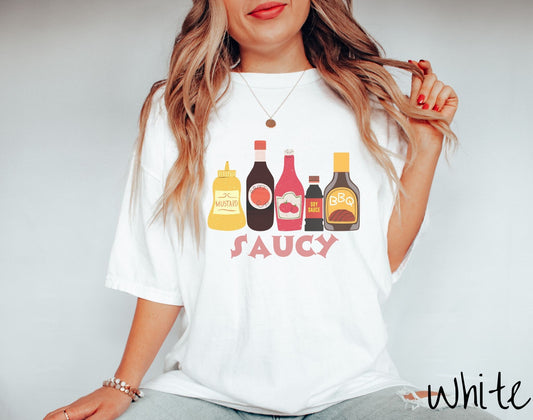 A woman wearing a vintage, white colored shirt with a mustard bottle, ketchup bottle, soy sauce bottle, barbecue sauce bottle, and another sauce bottle centered on the shirt with the text Saucy underneath the bottles.