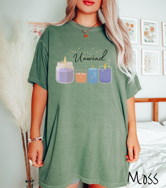 A woman wearing a vintage, moss colored shirt with the text Unwind at the top with twinkling, golden stars behind it, and below the text are a lit purple candle, a lit orange candle, a lit blue candle, and a lit grape candle.