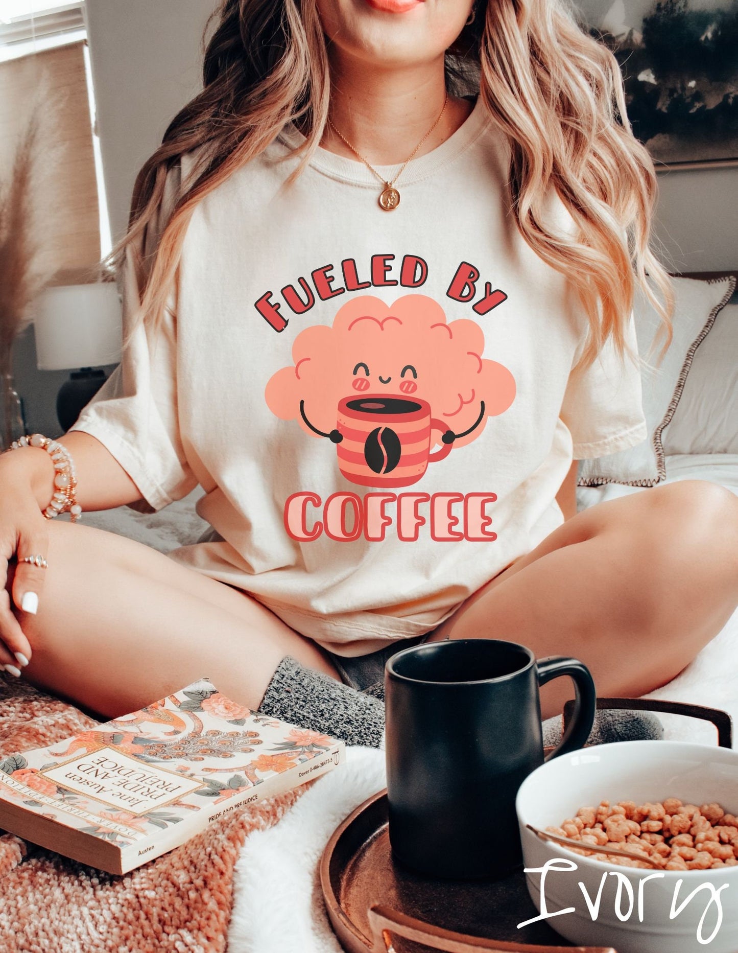 A woman wearing a cute ivory colored shirt with the text Fueled by Coffee and a puffy cloud smiling holding a cup of coffee.