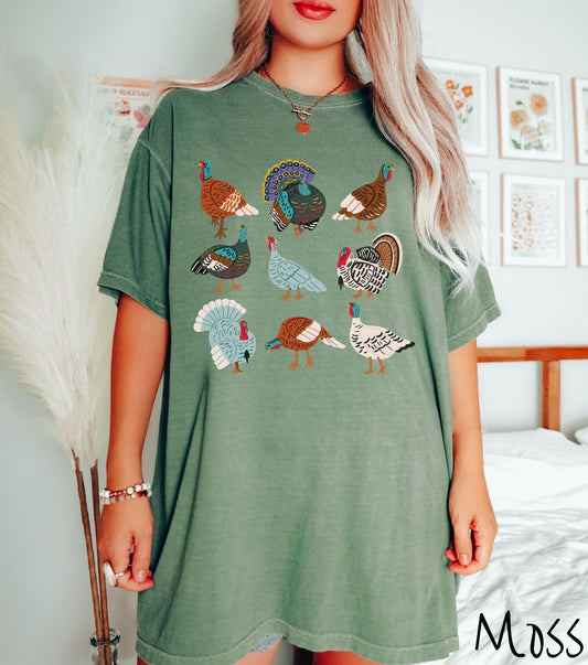 A woman wearing a cute, vintage moss colored Comfort Colors t-shirt with a three by three grid of different colored turkeys and holiday birds. Some are white, others brown and light blue, and all have red wattles.