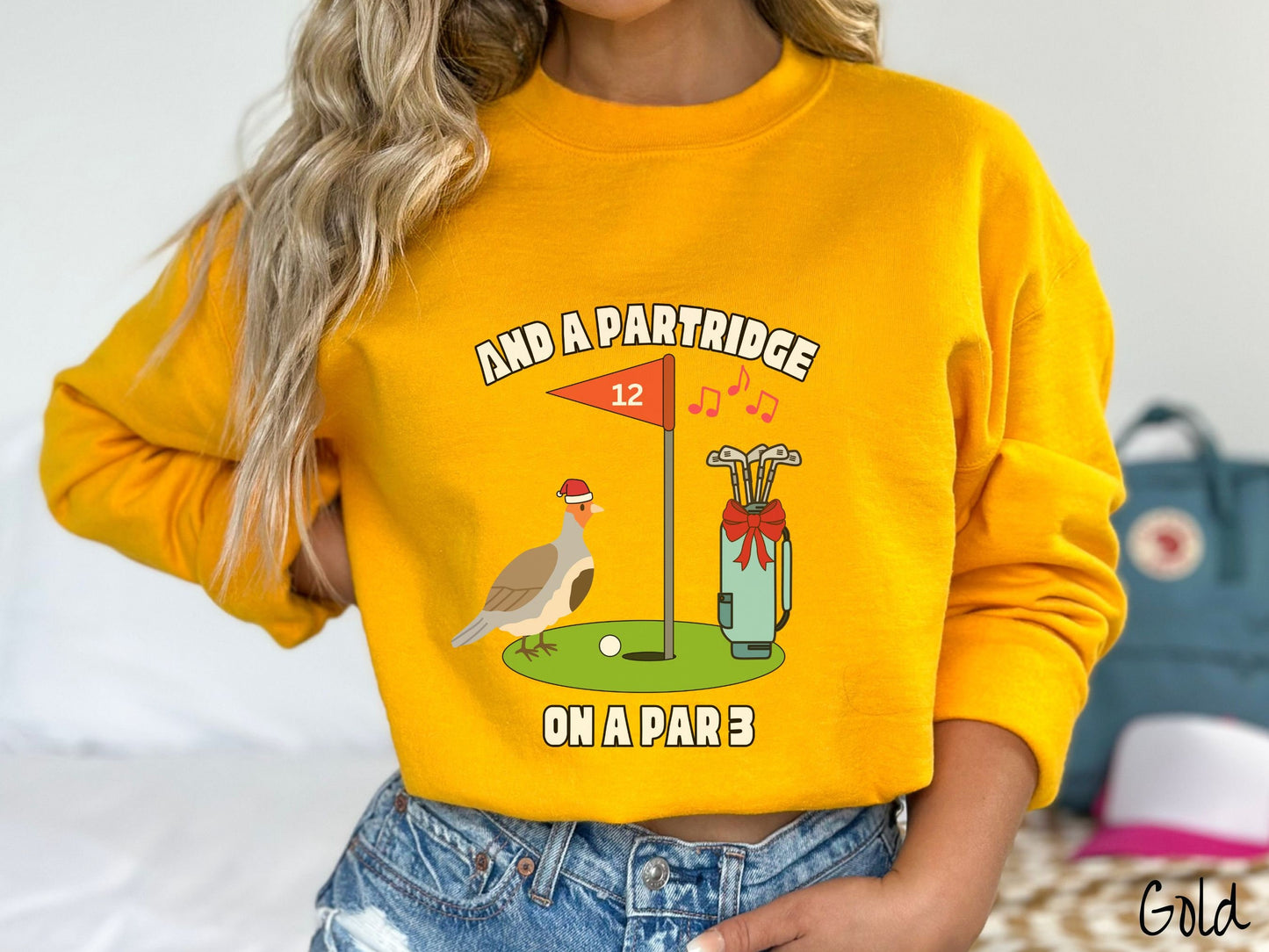 A woman wearing a cute gold colored sweatshirt with a partridge bird on a golf green next to a hole with a flag in it with #12 on it. There is also a set of golf clubs with a red bow and music playing in the air.