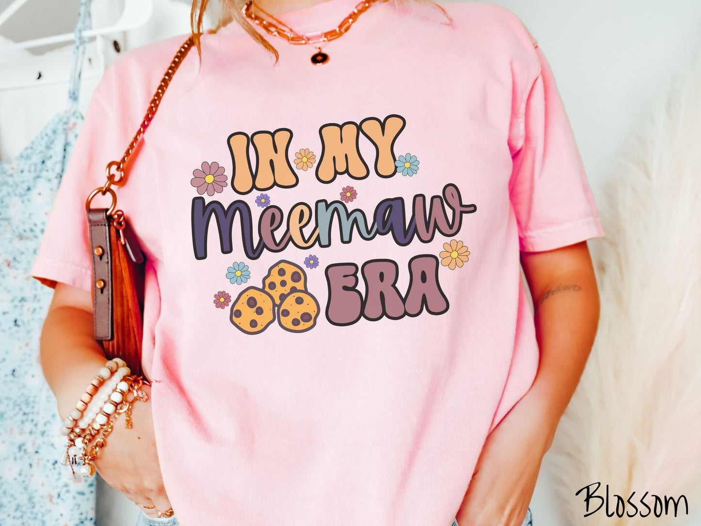 A woman wearing a cute blossom colored sweatshirt with text on the front saying In My Meemaw Era, with flowers and chocolate chip cookies mixed within the text.