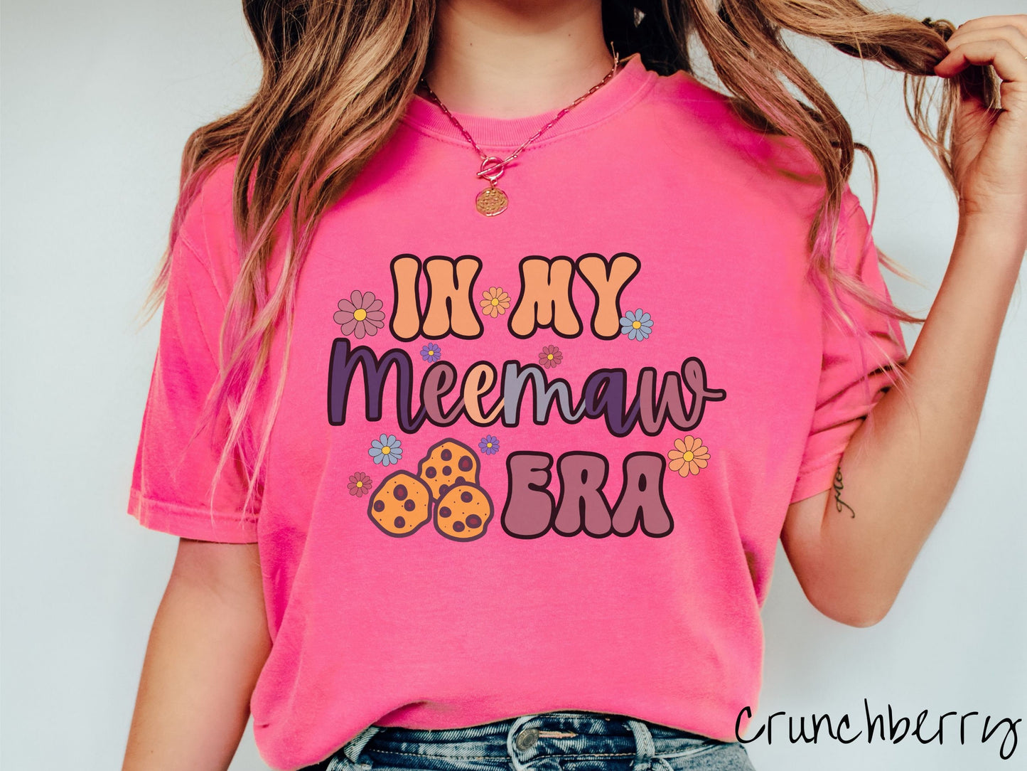 A woman wearing a cute crunchbery colored sweatshirt with text on the front saying In My Meemaw Era, with flowers and chocolate chip cookies mixed within the text.