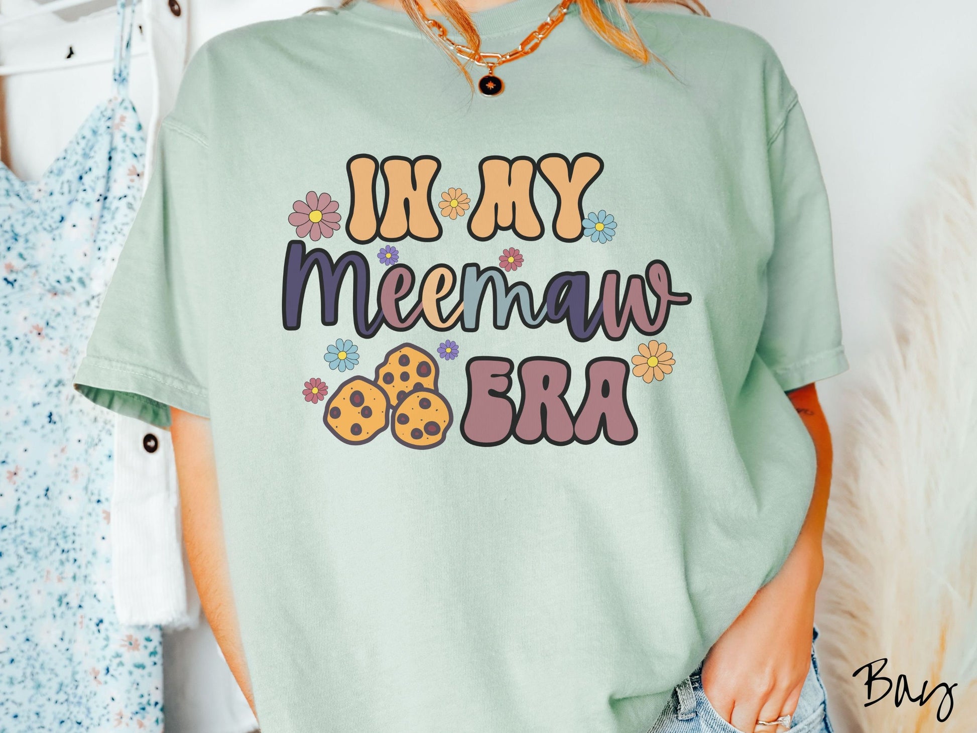 A woman wearing a cute bay colored sweatshirt with text on the front saying In My Meemaw Era, with flowers and chocolate chip cookies mixed within the text.