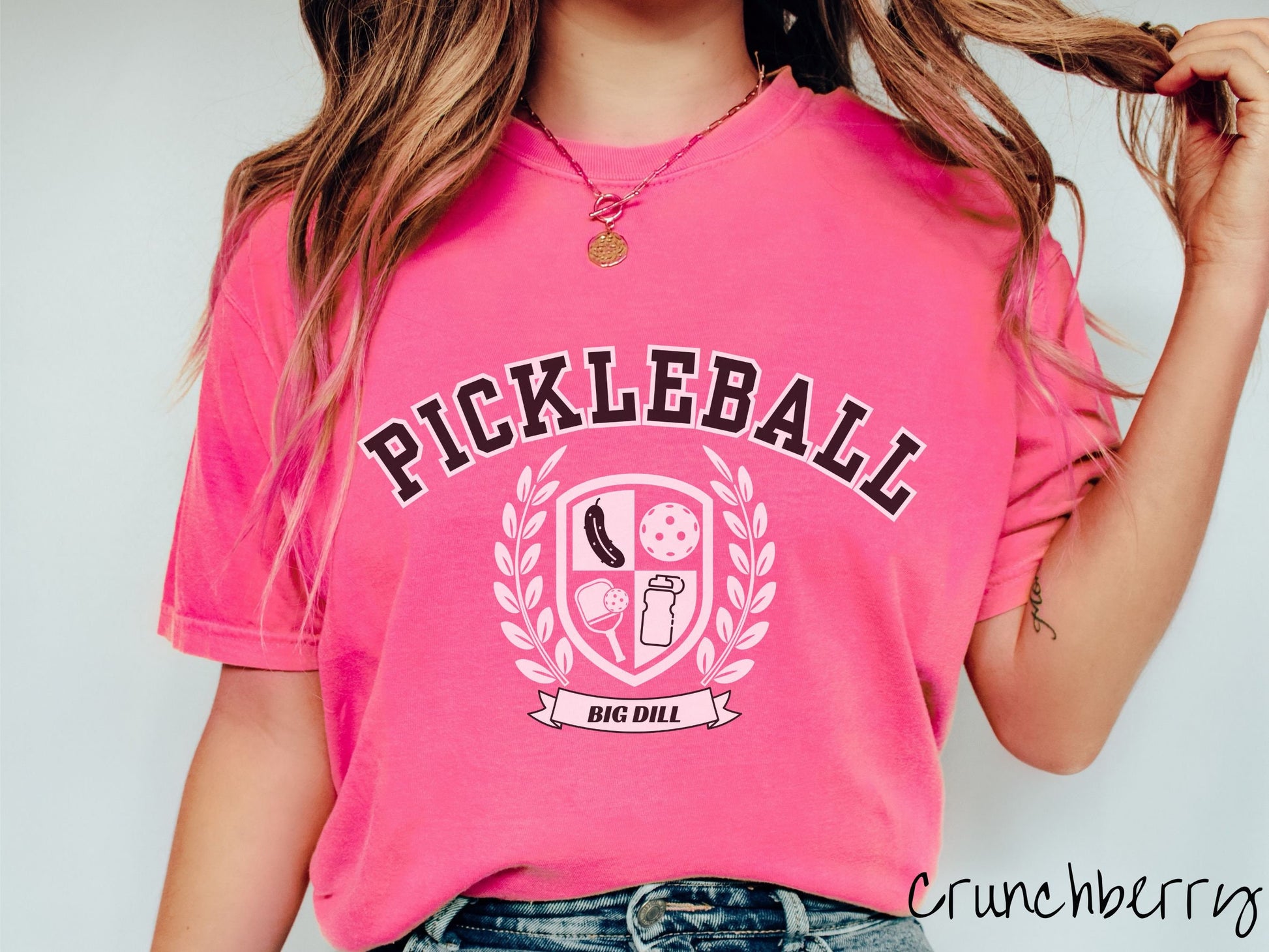 A woman wearing a cute crunchberry colored shirt with the text Pickleball across the top, a varsity logo centered underneath, and the words Big Dill underneath the logo.
