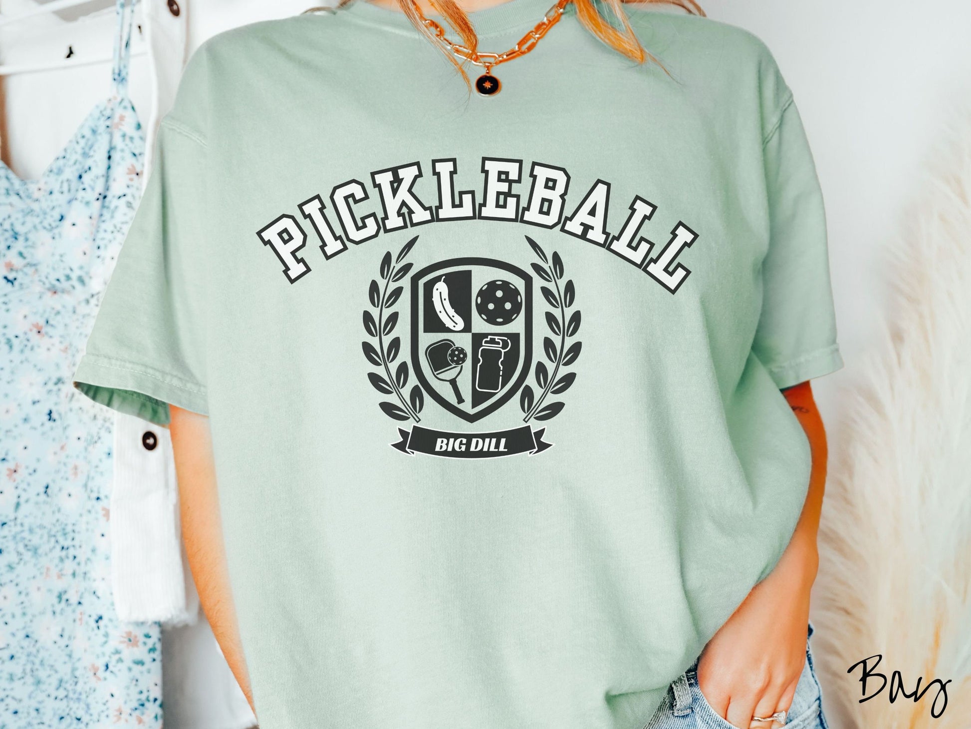 A woman wearing a cute bay colored shirt with the text Pickleball across the top, a varsity logo centered underneath, and the words Big Dill underneath the logo.