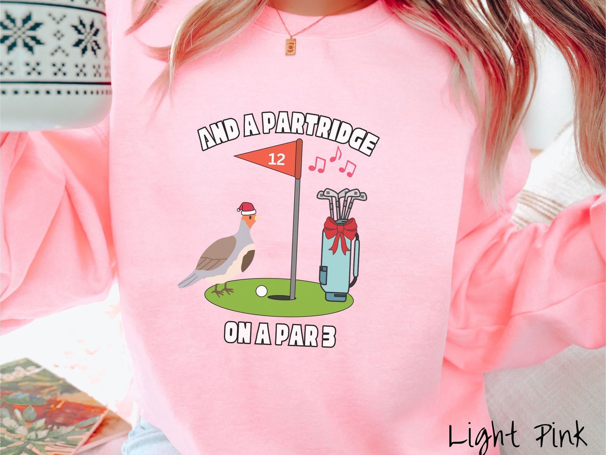A woman wearing a cute light pink colored sweatshirt with a partridge bird on a golf green next to a hole with a flag in it with #12 on it. There is also a set of golf clubs with a red bow and music playing in the air.