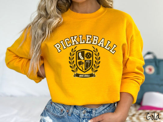 A woman wearing a cute gold colored sweatshirt with the text Pickleball across the top, a varsity logo centered underneath, and the words Big Dill underneath the logo.