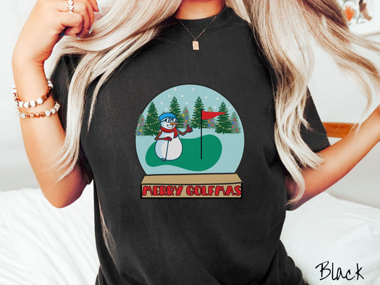 A woman wearing a cute pepper colored shirt with red text saying Merry Golfmas and a picture of a snowman giving the thumbs up on the golf green next to the hole with a flag in it.