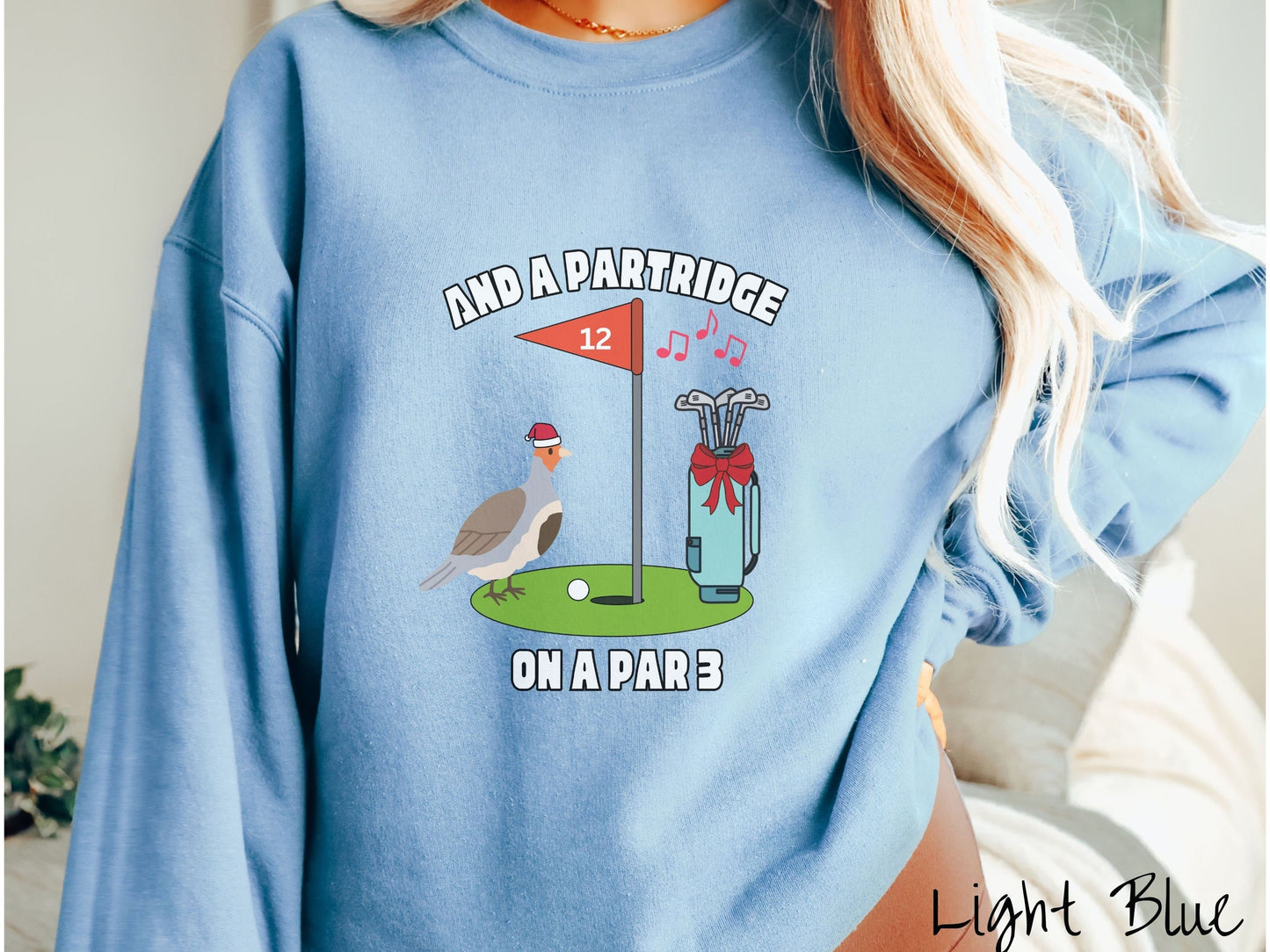A woman wearing a cute light blue colored sweatshirt with a partridge bird on a golf green next to a hole with a flag in it with #12 on it. There is also a set of golf clubs with a red bow and music playing in the air.