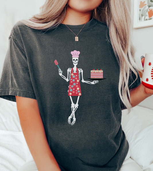 A woman wearing a cute black colored shirt with a female skeleton wearing a pink chef’s hat standing cross legged in a red apron adorned with flowers holding a cake on a plate in her left hand and a spatula in her right hand.