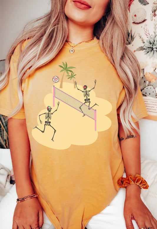 A woman wearing a cute mustard colored shirt with two skeletons playing sand volleyball over a pink net with palm trees in the background.