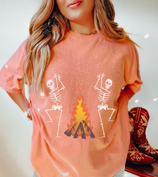 A woman wearing a cute terracotta colored shirt with two white skeletons dancing and clapping hands around a roaring bonfire.