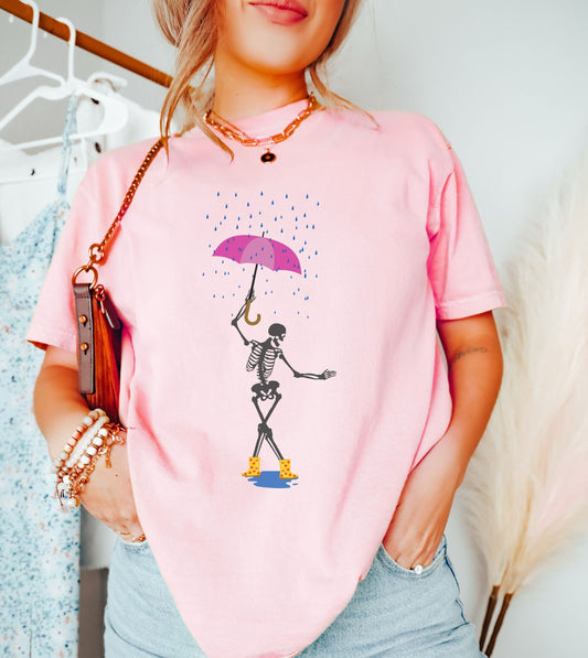 A woman wearing a cute pink colored shirt with a female skeleton dancing in the rain with a pink and purple umbrella and wearing yellow with red polka dot rain boots.