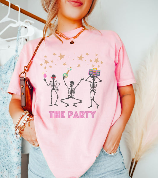 A woman wearing a cute pink colored shirt with three skeletons holding alcoholic drinks, liquor, and boomboxes dancing under a sky of stars with the text The Party written underneath the skeletons.