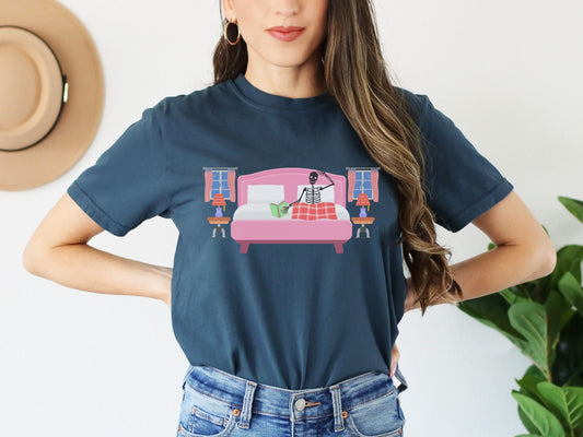 A woman wearing a cute midnight colored shirt with a skeleton lying in a pink bed covered with a red blanket reading a green book, two night tables and windows on each side of the bed.