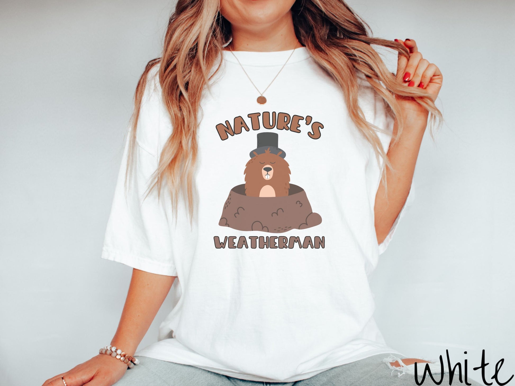 A woman wearing a cute white colored shirt with the text Natures Weatherman sandwiching Phil the Punxsutawney PA groundhog who is wearing a black top hat and is coming out of his hole for Groundhogs Day.