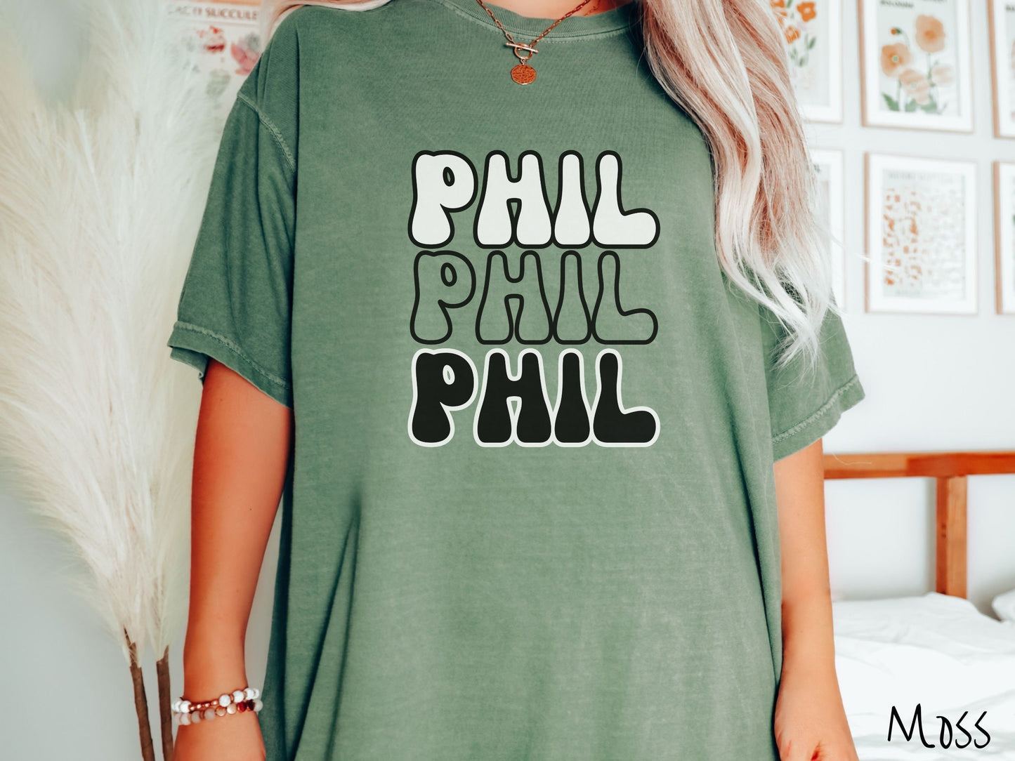 A woman wearing a cute moss colored shirt with the text PHIL listed three times vertically in white, red, and black color text. The shirt is referring to the chant that occurs for the groundhog Phil every Groundhogs Day in Punxsutawney, PA.