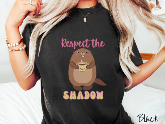 A woman wearing a cute black colored shirt with the pink text Respect the Shadow sandwiching a pink cheeked groundhog holding a steaming cup of coffee that has a smiling face on the front and the groundhog is displaying a look of sheer joy.