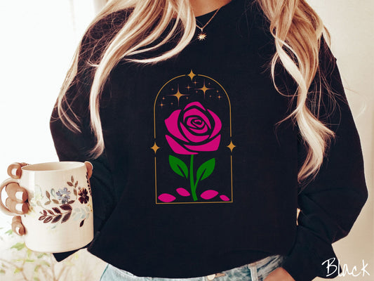 A woman wearing a cute, vintage black colored sweatshirt with an enchanted pink rose in a golden clear container with gold stars twinkling above and pink flower petals resting below.