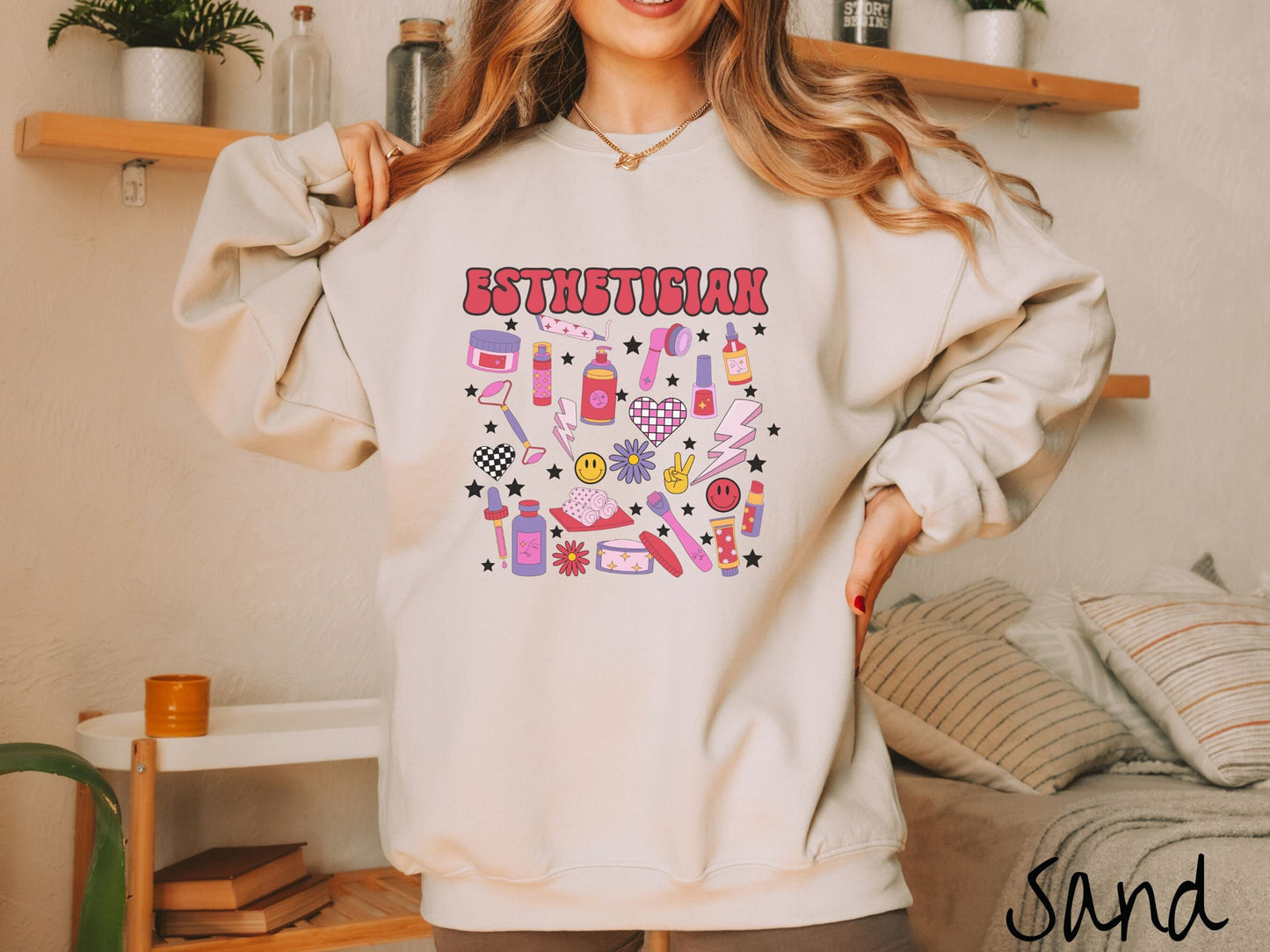 A woman wearing a cute sand colored sweatshirt with the word Esthetician across the top and hair care items below like cream, rollers, hair care products, and droppers. Smiley faces, stars, and lightning bolts are spread out among the items.