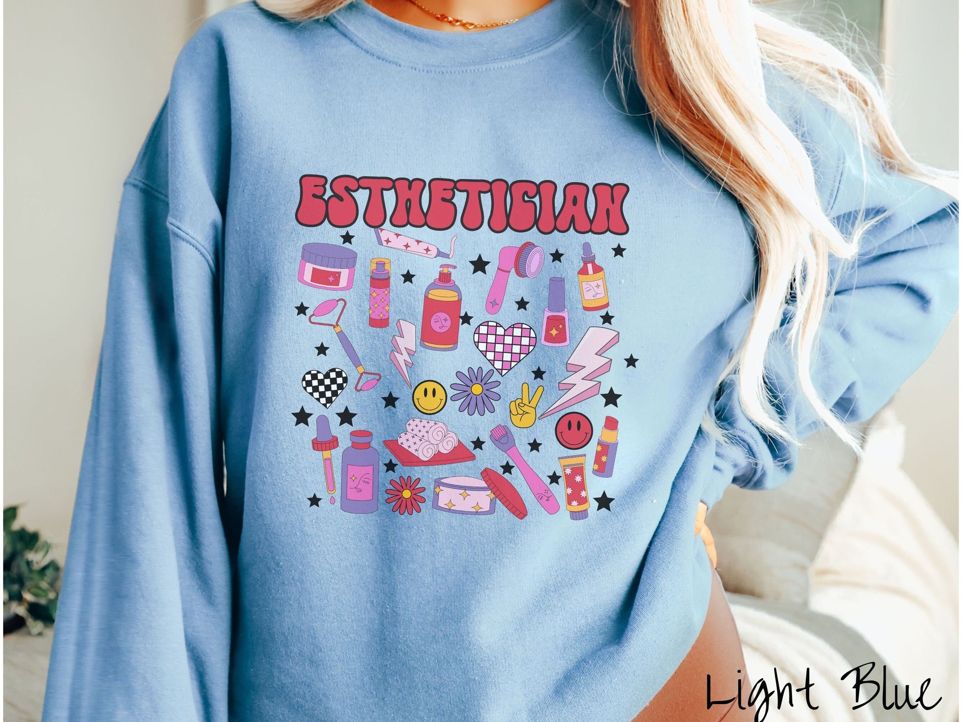 A woman wearing a cute light blue colored sweatshirt with the word Esthetician across the top and hair care items below like cream, rollers, hair care products, and droppers. Smiley faces, stars, and lightning bolts are spread out among the items.