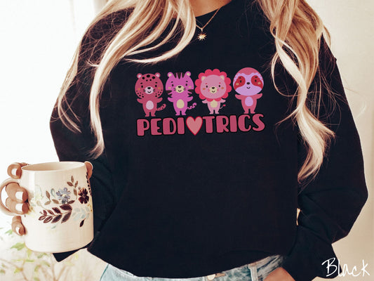 A woman wearing a cute black colored sweatshirt with the text Pediatrics in the center, a heart replacing the letter A, and above the text are a maroon lion, a pink tiger, a red lion, and a pink sloth all with their hands open as if asking for a hug.