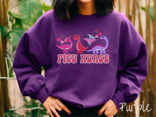 A woman wearing a cute purple colored sweatshirt with the text PICU Nurse in the center below four dinosaurs, a pink Parasaurolophus, a red tyrannosaurus rex, a green pterodactyl, a purple Brachiosaurus, and pink and red hearts in the air.