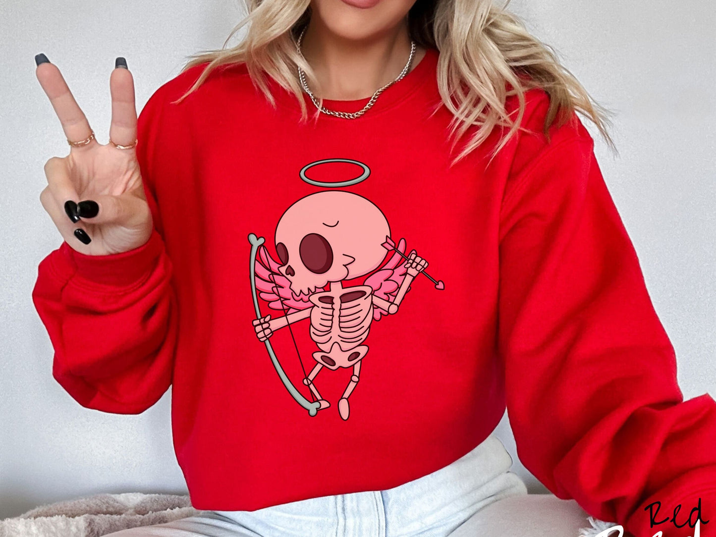 A woman wearing a cute red colored sweatshirt with a pink winged baby skeleton cupid with a halo holding a heart-tipped arrow in its left hand and a bow in its right hand.