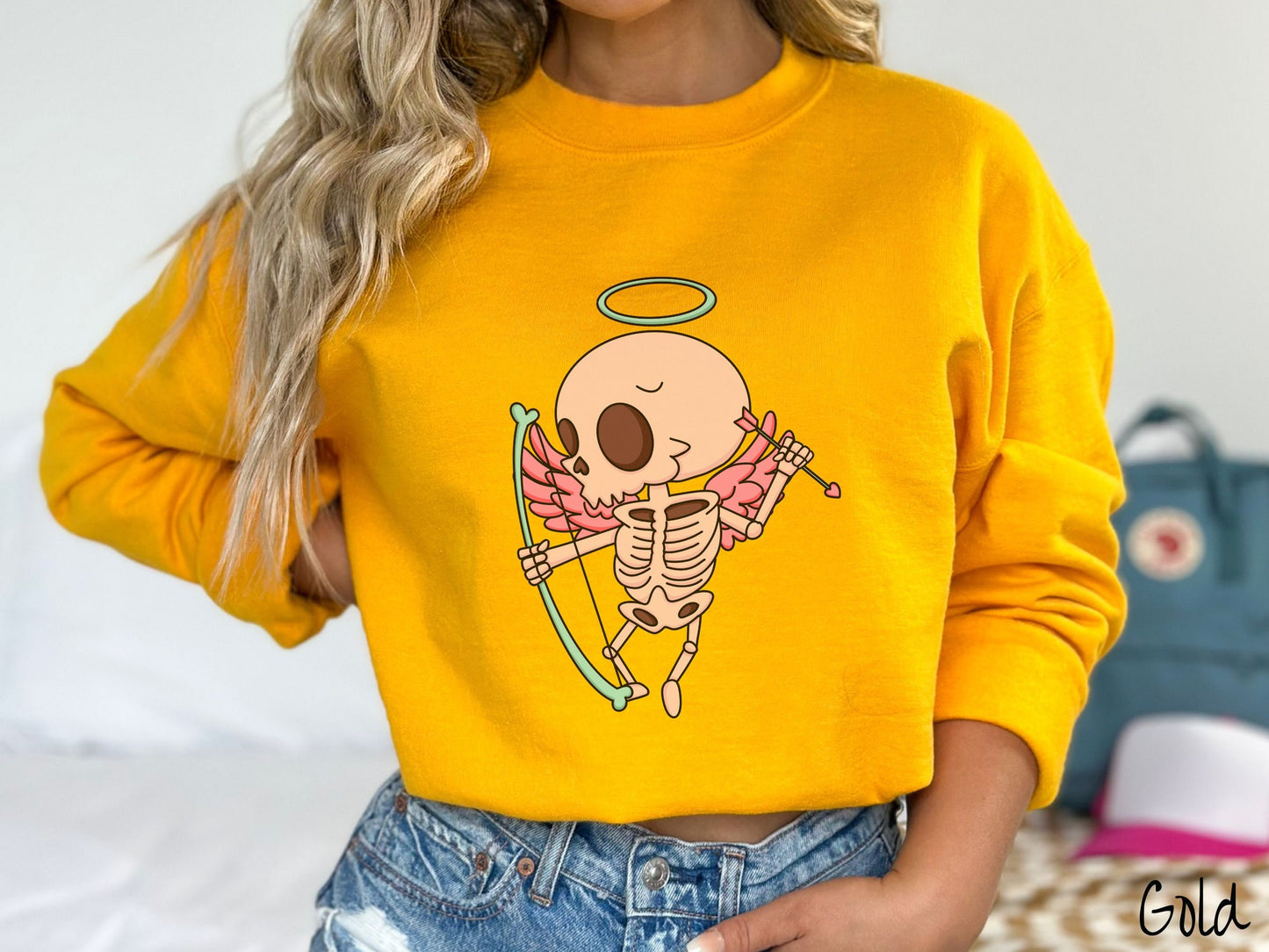 A woman wearing a cute gold colored sweatshirt with a pink winged baby skeleton cupid with a halo holding a heart-tipped arrow in its left hand and a bow in its right hand.
