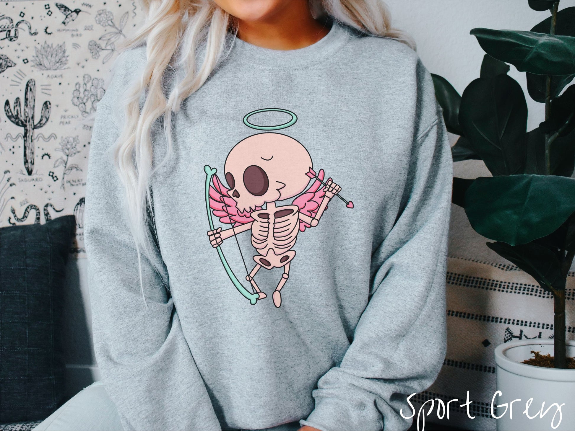 A woman wearing a cute sport grey colored sweatshirt with a pink winged baby skeleton cupid with a halo holding a heart-tipped arrow in its left hand and a bow in its right hand.
