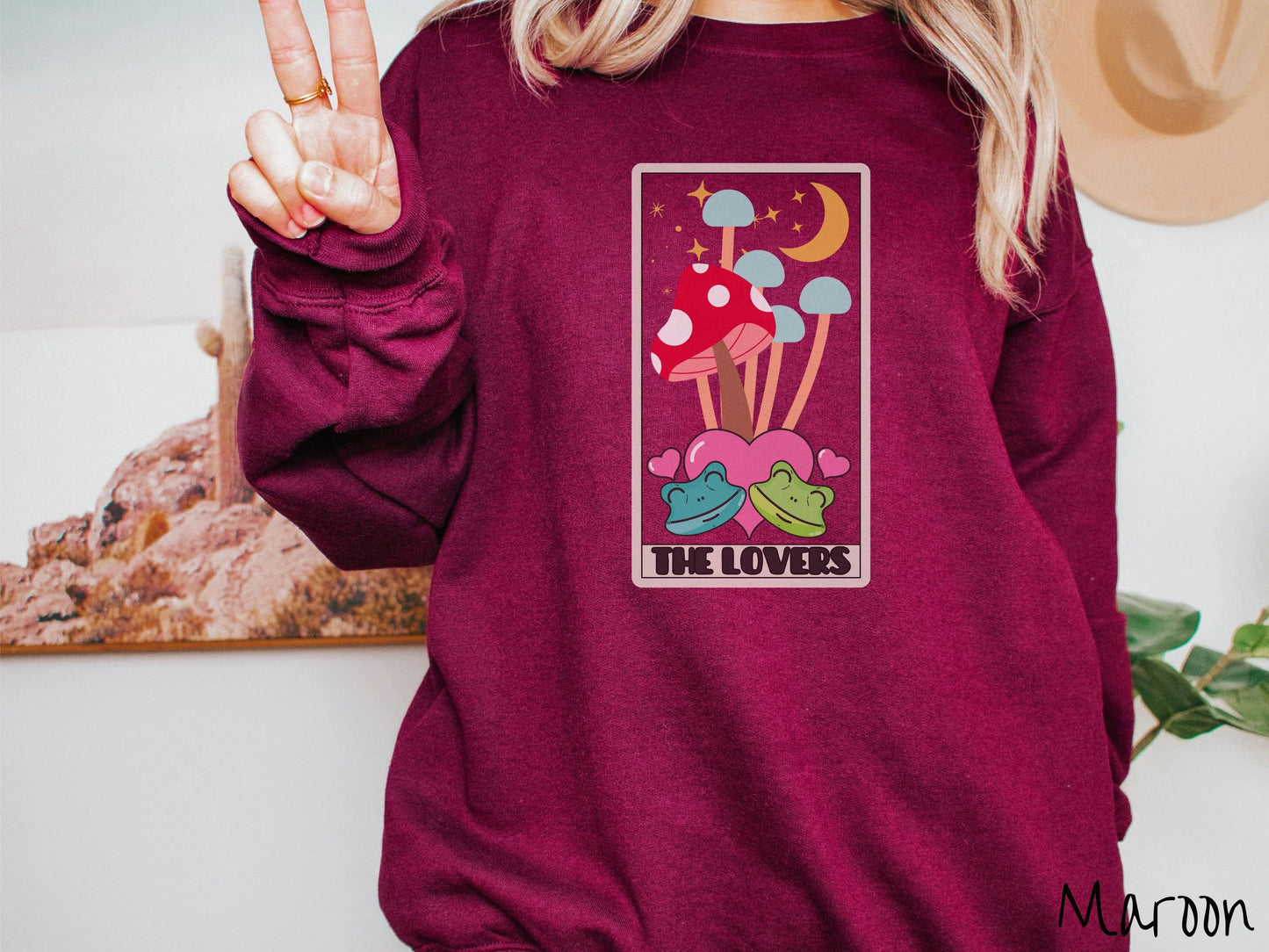 A woman wearing a cute maroon colored sweatshirt with a large red mushroom and 4 turquoise mushrooms in the background, all under a crescent yellow moon and yellow stars, two green frogs smiling in front of pink hearts below the mushrooms.