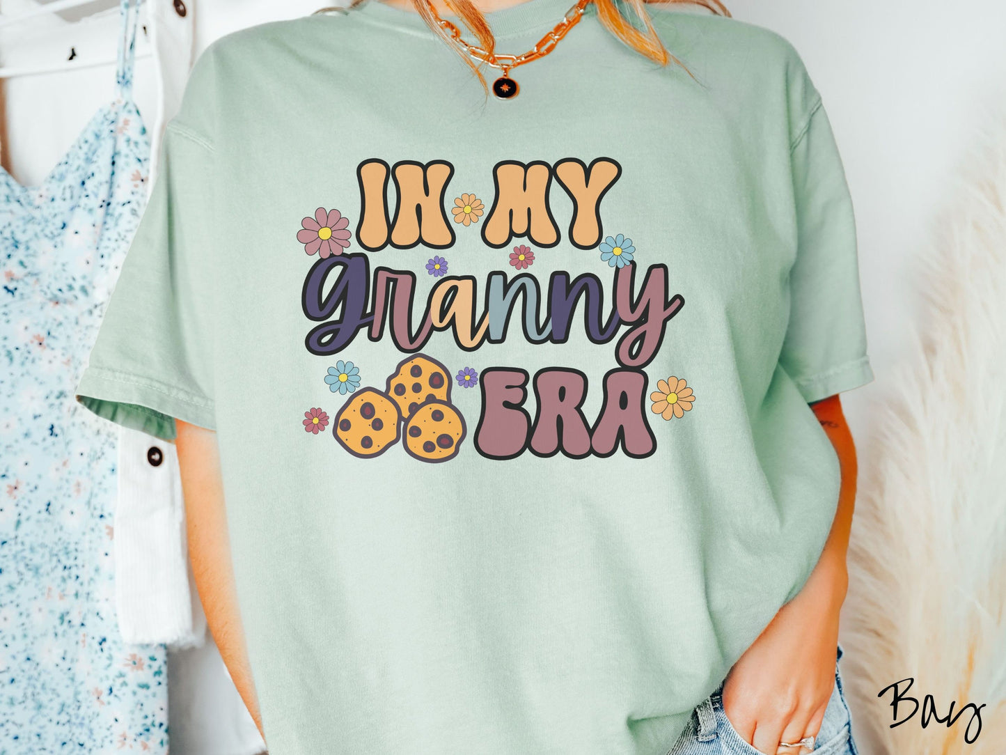 A woman wearing a cute bay colored sweatshirt with text on the front saying In My Granny Era, with flowers and chocolate chip cookies mixed within the text.
