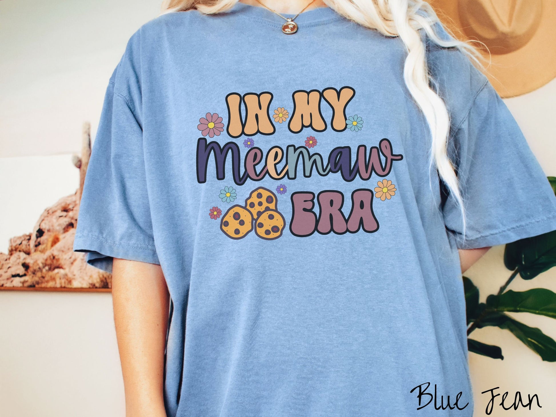 A woman wearing a cute blue jean colored sweatshirt with text on the front saying In My Meemaw Era, with flowers and chocolate chip cookies mixed within the text.