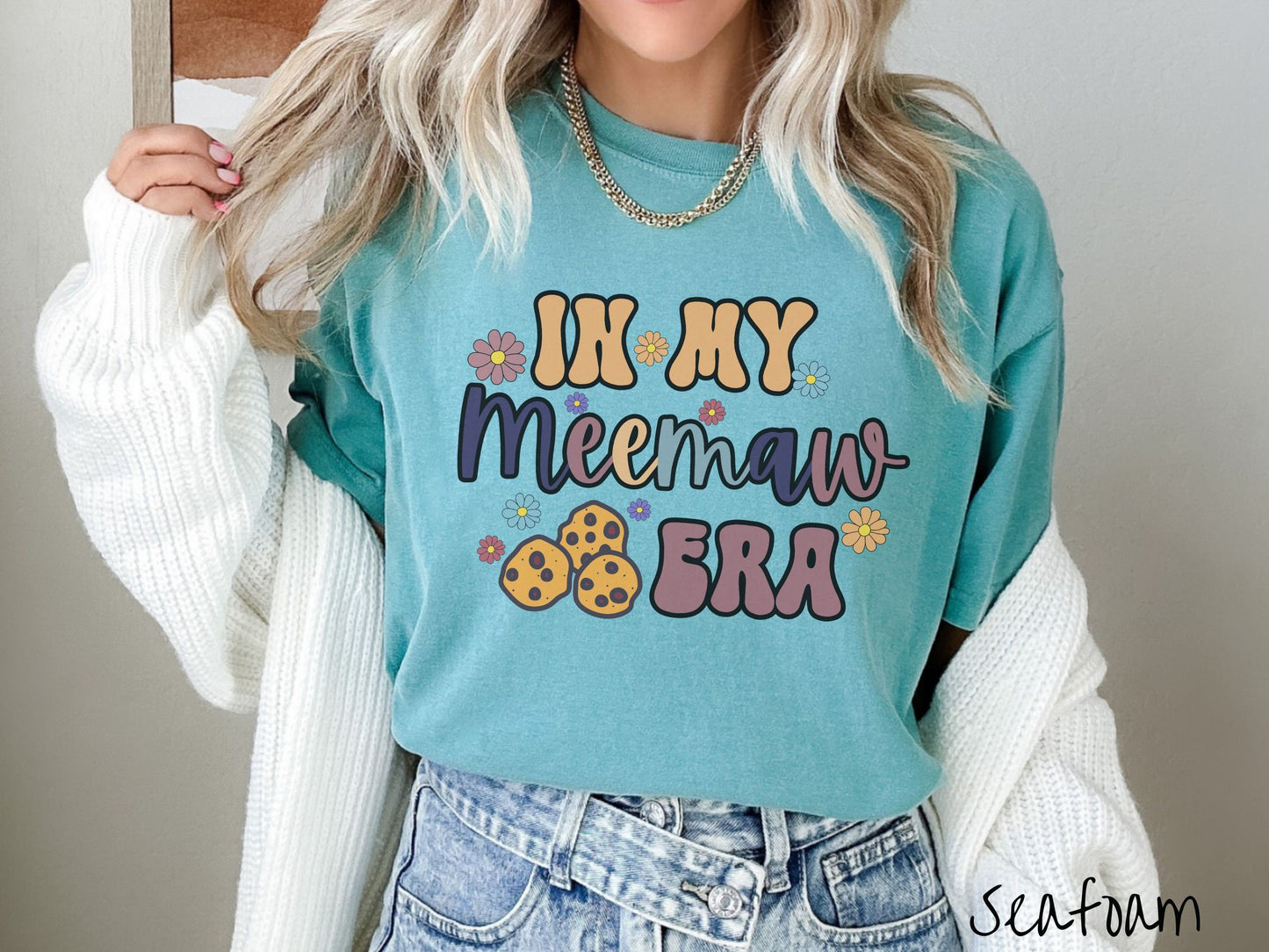 A woman wearing a cute seafoam colored sweatshirt with text on the front saying In My Meemaw Era, with flowers and chocolate chip cookies mixed within the text.