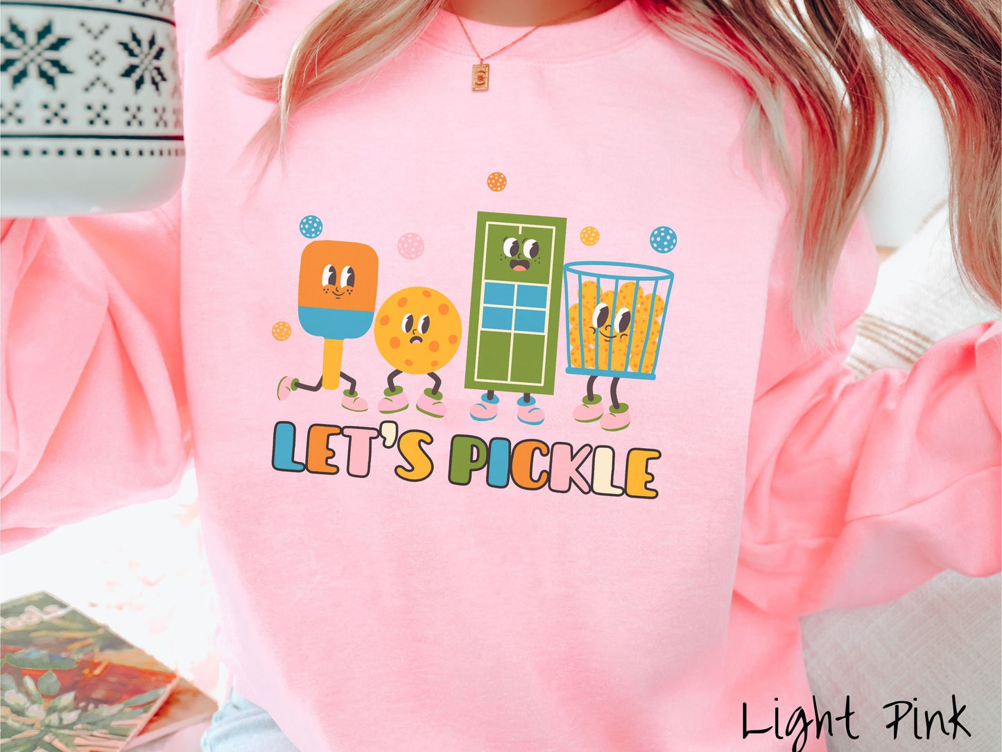 A woman wearing a cute light pink colored sweatshirt with the text Lets Pickle, and above the text are a popsicle, cookie, popcorn, and pickle balls.