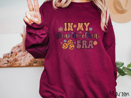 A woman wearing a cute maroon colored sweatshirt with text on the front saying In My Grandmother Era, with flowers and chocolate chip cookies mixed within the text.