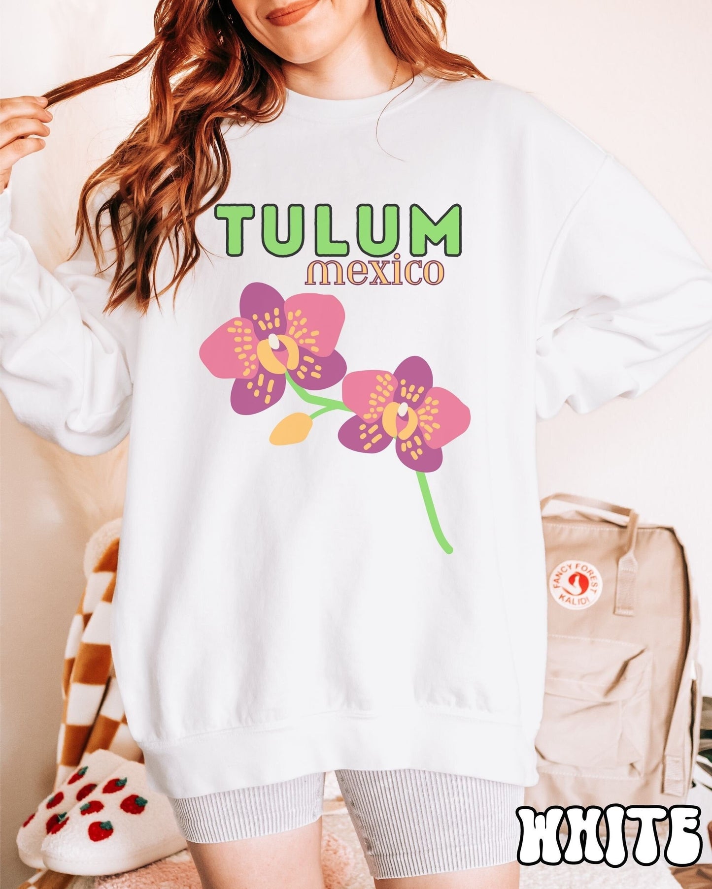 A woman wearing a vintage, pepper colored sweatshirt with the text Tulum Mexico in green and yellow font, respectively, at the top and below it are two beautiful pink and purple flowers with green stems.